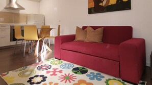 A living room with a red sofa, a colorful floral rug, a painting of a sunset, and a kitchen area with a stove and chairs. - Casa Cadu - Porto (Porto)