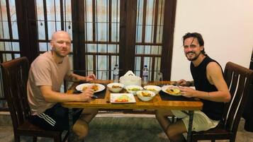 Twee personen zitten aan een houten tafel met verschillende gerechten, in een kamer met grote ramen en houten deuren.