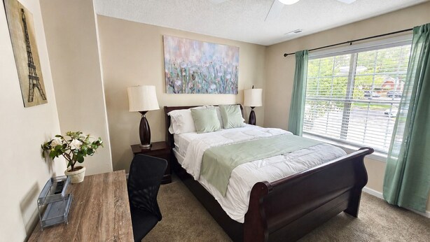 A hotel room with a bed, two bedside lamps, a desk with a chair, a potted plant, and a painting on the wall. - 3BR Retreat • Perfect for Groups • Near Downtown (Raleigh)