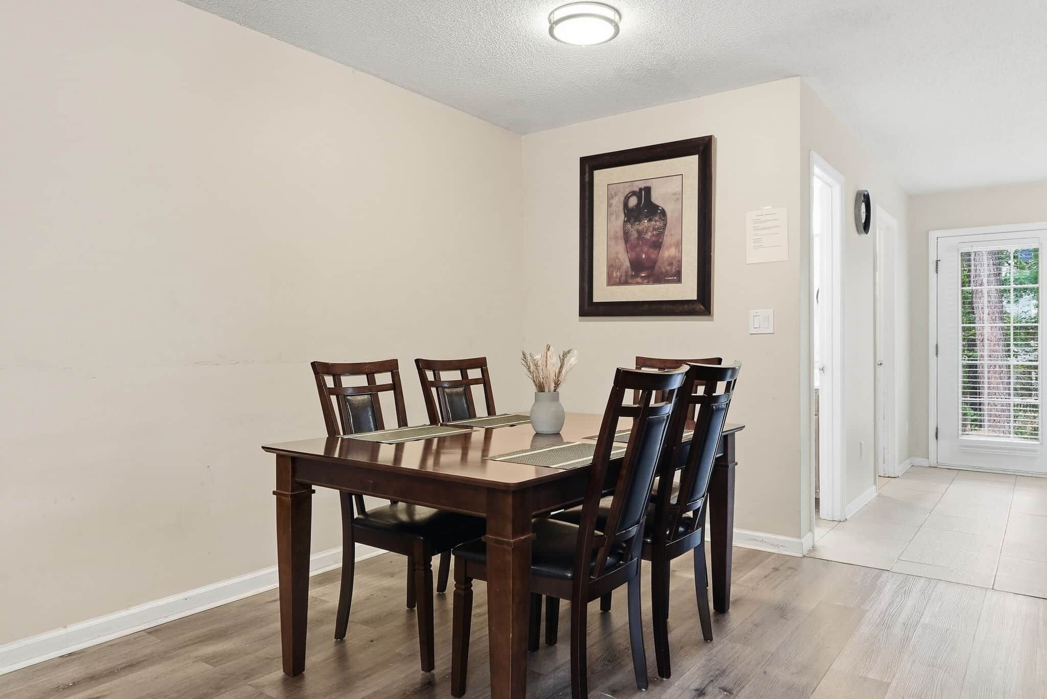 Un comedor con una mesa y sillas de madera, un jarrón con flores secas y un cuadro en la pared.