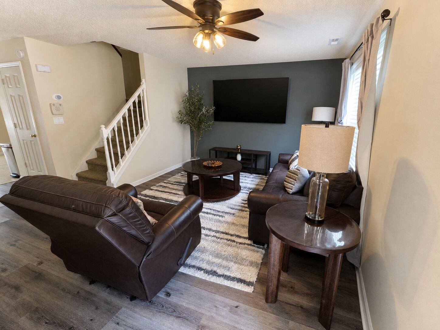 A living room with a brown leather sofa, a coffee table, a TV mounted on the wall, and a ceiling fan.