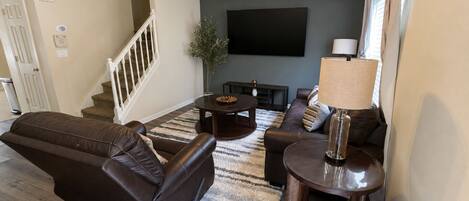 A living room with a brown leather sofa, a coffee table, a TV mounted on the wall, and a ceiling fan.