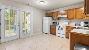 A kitchen with white appliances, wooden cabinets, a double door with blinds, and a tiled floor. - 3BR Retreat • Perfect for Groups • Near Downtown (Raleigh)