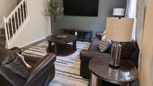 A living room with a dark brown leather sofa, a round coffee table, a TV mounted on the wall, and a staircase with white railings. - 3BR Retreat • Perfect for Groups • Near Downtown (Raleigh)