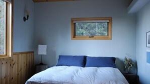 Un dormitorio con una cama de madera, una ventana con vistas a árboles, una lámpara de noche y un cuadro enmarcado en la pared.