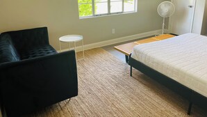 A bedroom with a bed, a black chair, a small white table, a standing fan, and a window with a view of greenery. - Quiet studio in New Port Richey Downtown. Apartment B (New Port Richey)