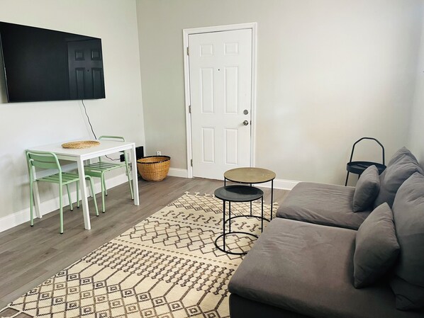 A living room with a sofa, a small table, a TV mounted on the wall, and a door.