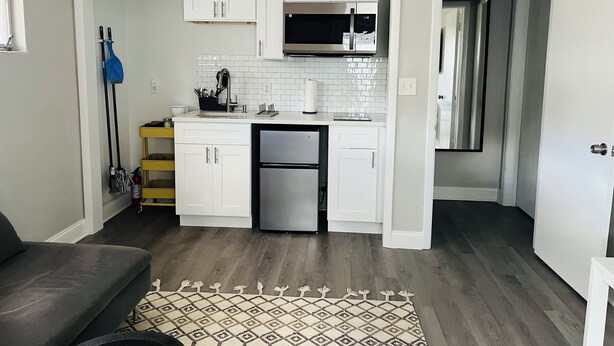 A compact kitchen with white cabinets, a stainless steel refrigerator, and a microwave. A patterned rug is on the floor, and a mop is leaning against the wall. - Quiet studio in New Port Richey Downtown. Apartment B (New Port Richey)