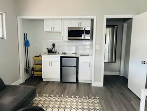 A compact kitchen with white cabinets, a stainless steel refrigerator, and a microwave. A patterned rug is on the floor, and a mop is leaning against the wall.