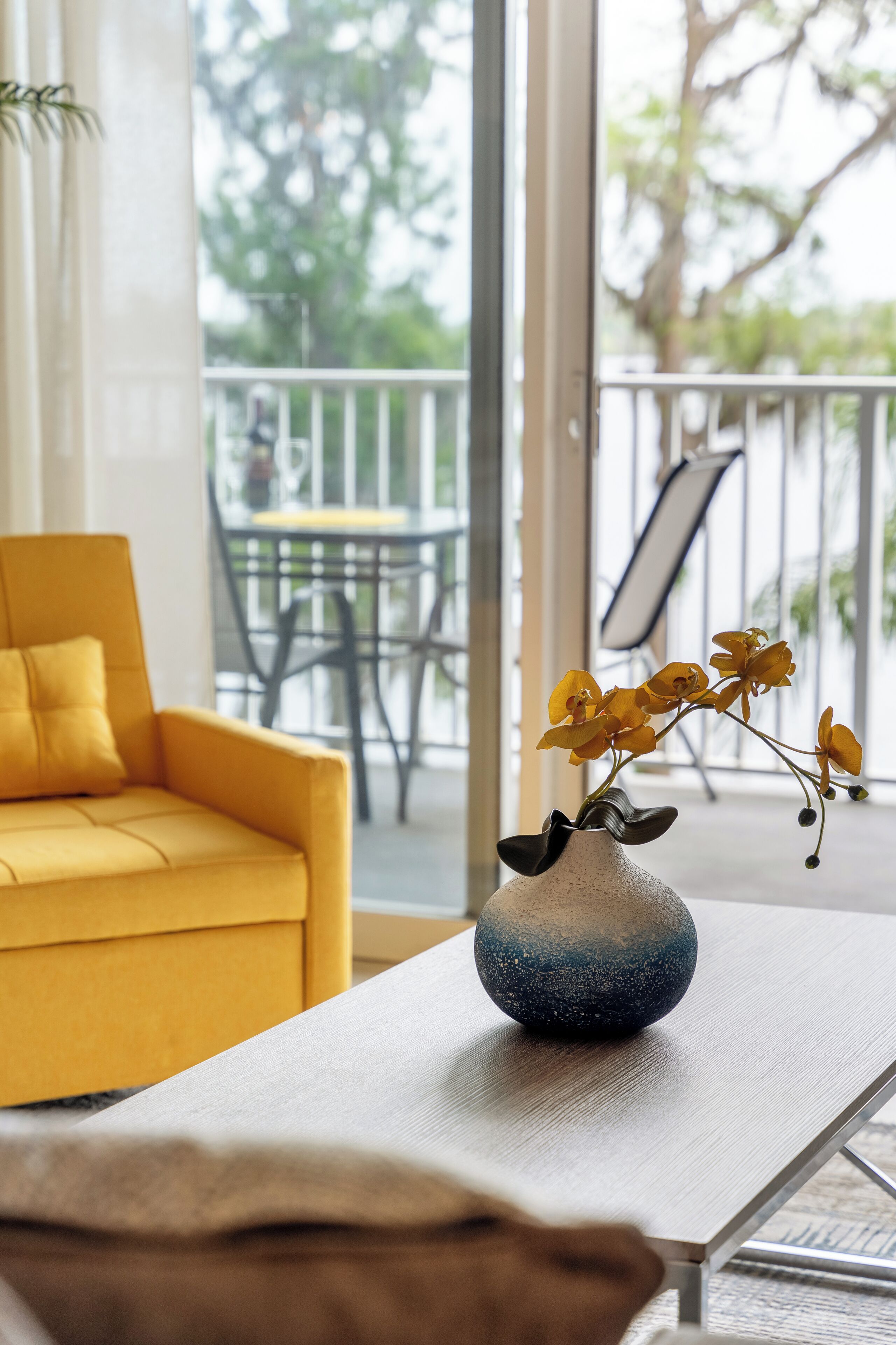 A yellow sofa, a white table, and a vase with yellow flowers on a balcony with a view of trees and a chair.