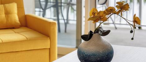A yellow sofa, a white table, and a vase with yellow flowers on a balcony with a view of trees and a chair.