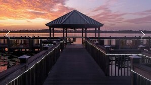 Une jetée en bois mène à un belvédère surmonté d’un toit en métal, d’où l’on peut admirer un plan d’eau au coucher du soleil.