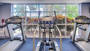 Une salle de sport équipée de tapis de course et d’un appareil elliptique, avec de grandes fenêtres donnant sur des arbres et des plantes.