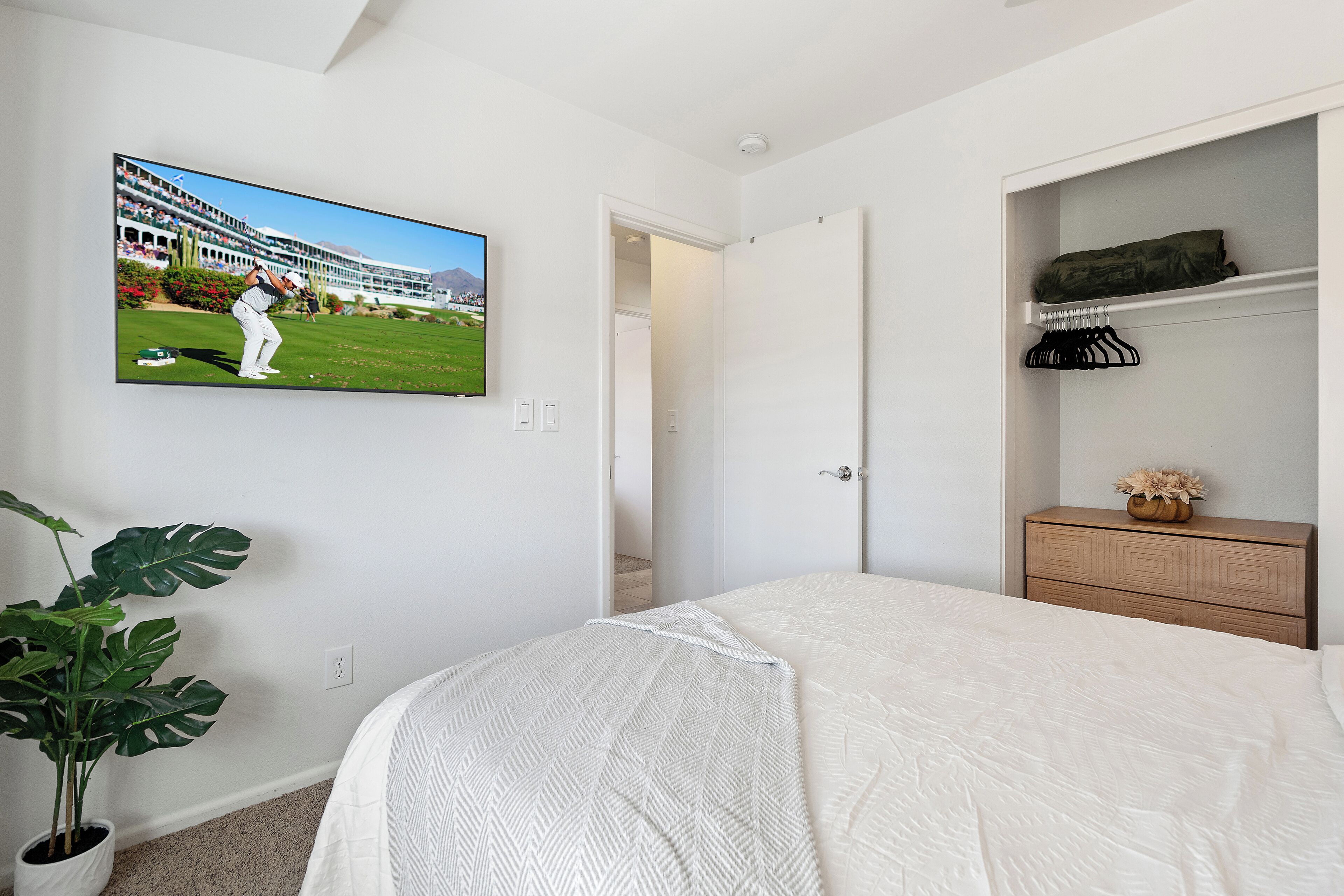 Una camera da letto con un letto, una TV a parete, una pianta in vaso e un comò di legno.