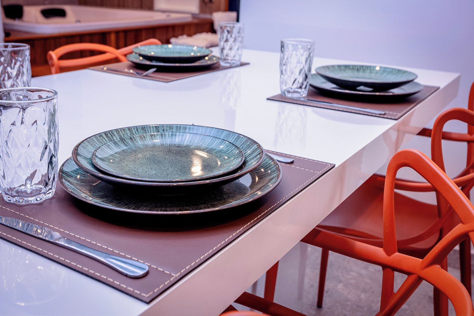 Una mesa de comedor preparada con platos, vasos y cubiertos, rodeada de sillas color naranja.