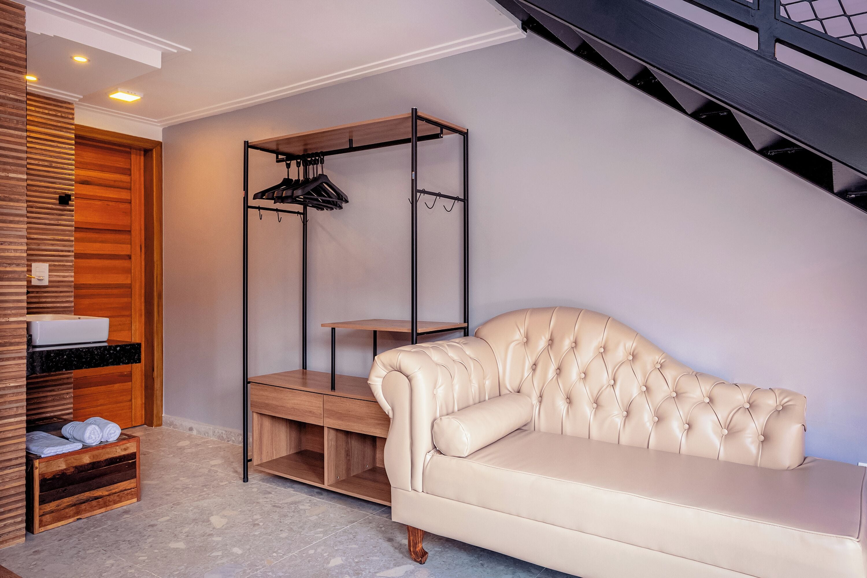 Een kamer met een beige leren bank, houten planken met handdoeken, een kapstok en een trap.