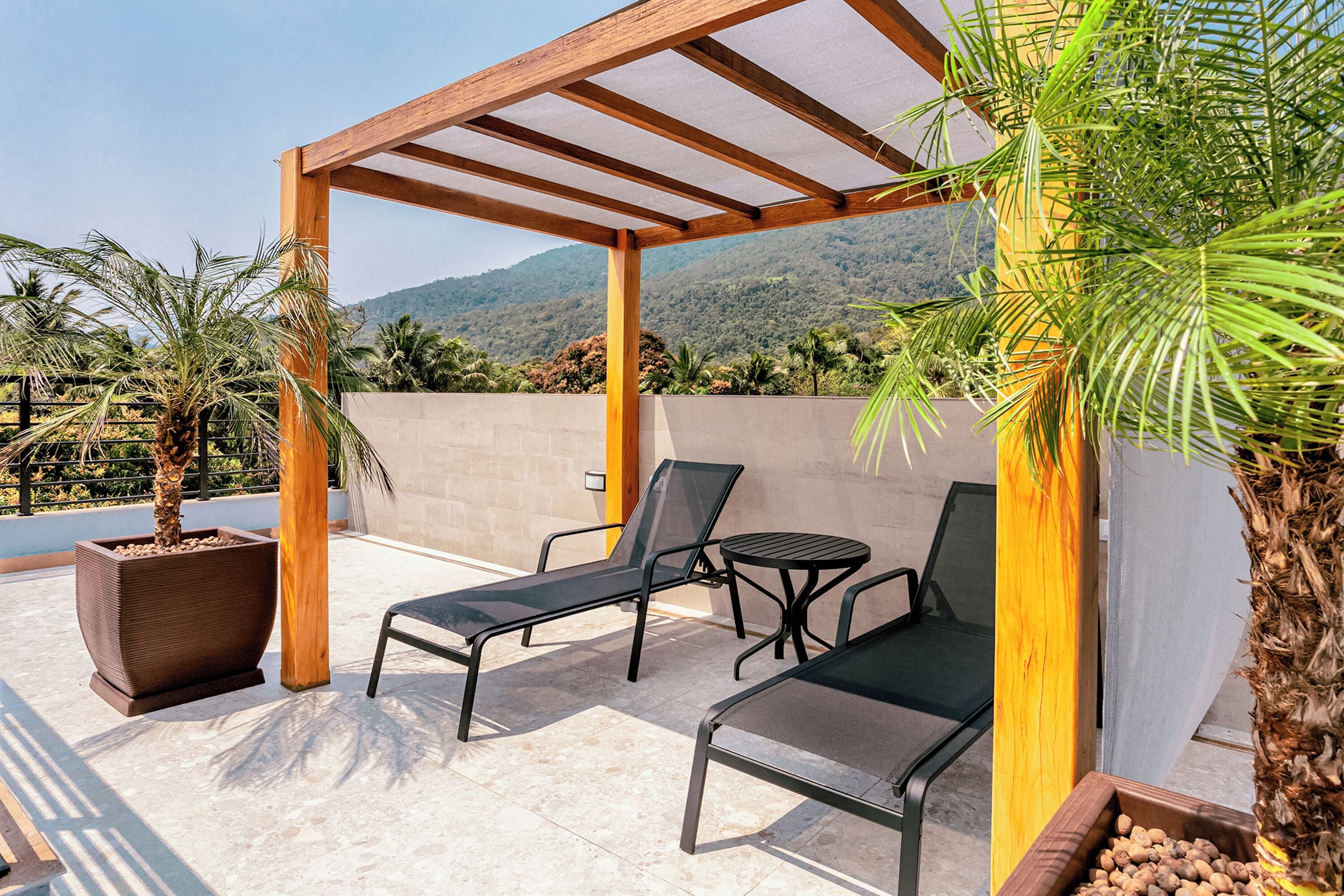 Um pátio externo com pérgola, duas poltronas de descanso, uma mesa pequena e plantas em vasos.