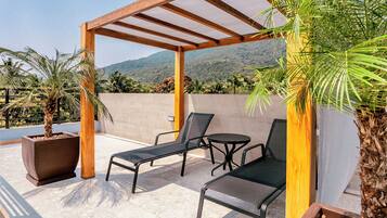 Um pátio externo com pérgola, duas poltronas de descanso, uma mesa pequena e plantas em vasos.