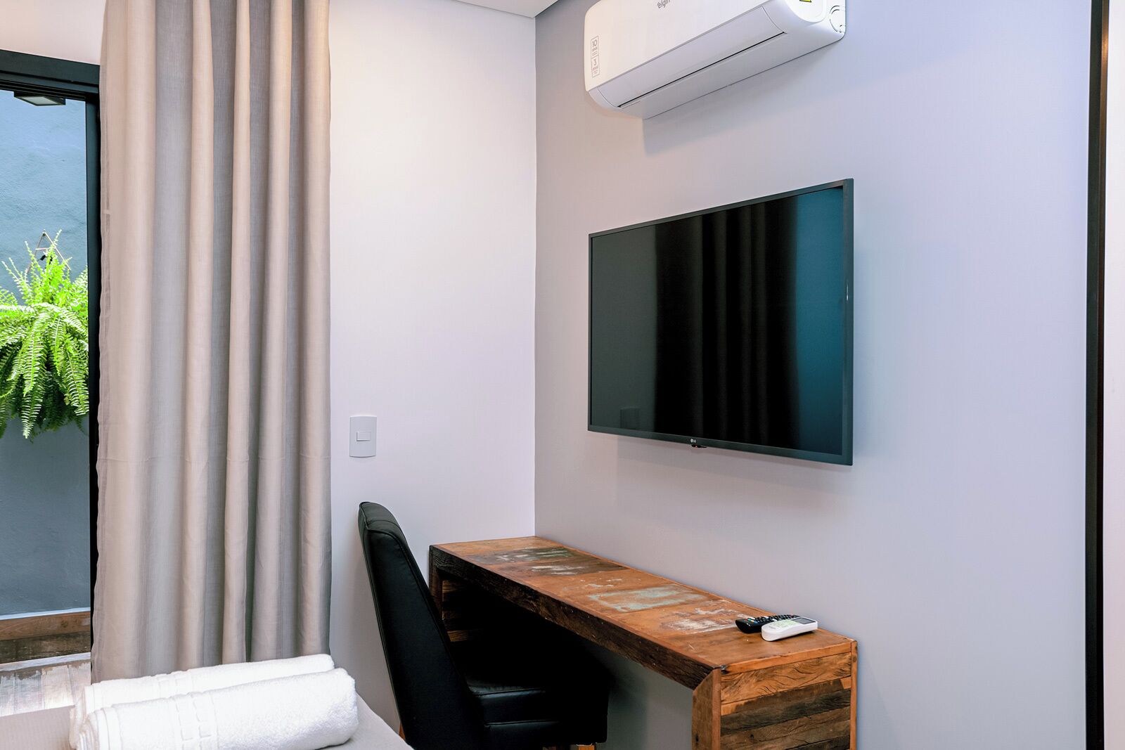 A modern room with a large window, a flat-screen TV mounted on the wall, a wooden desk, and a black chair.