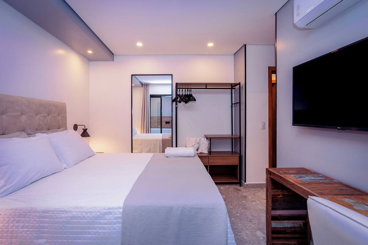 Quarto de hotel moderno com uma cama grande, um banco de madeira, uma TV e um closet espaçoso.