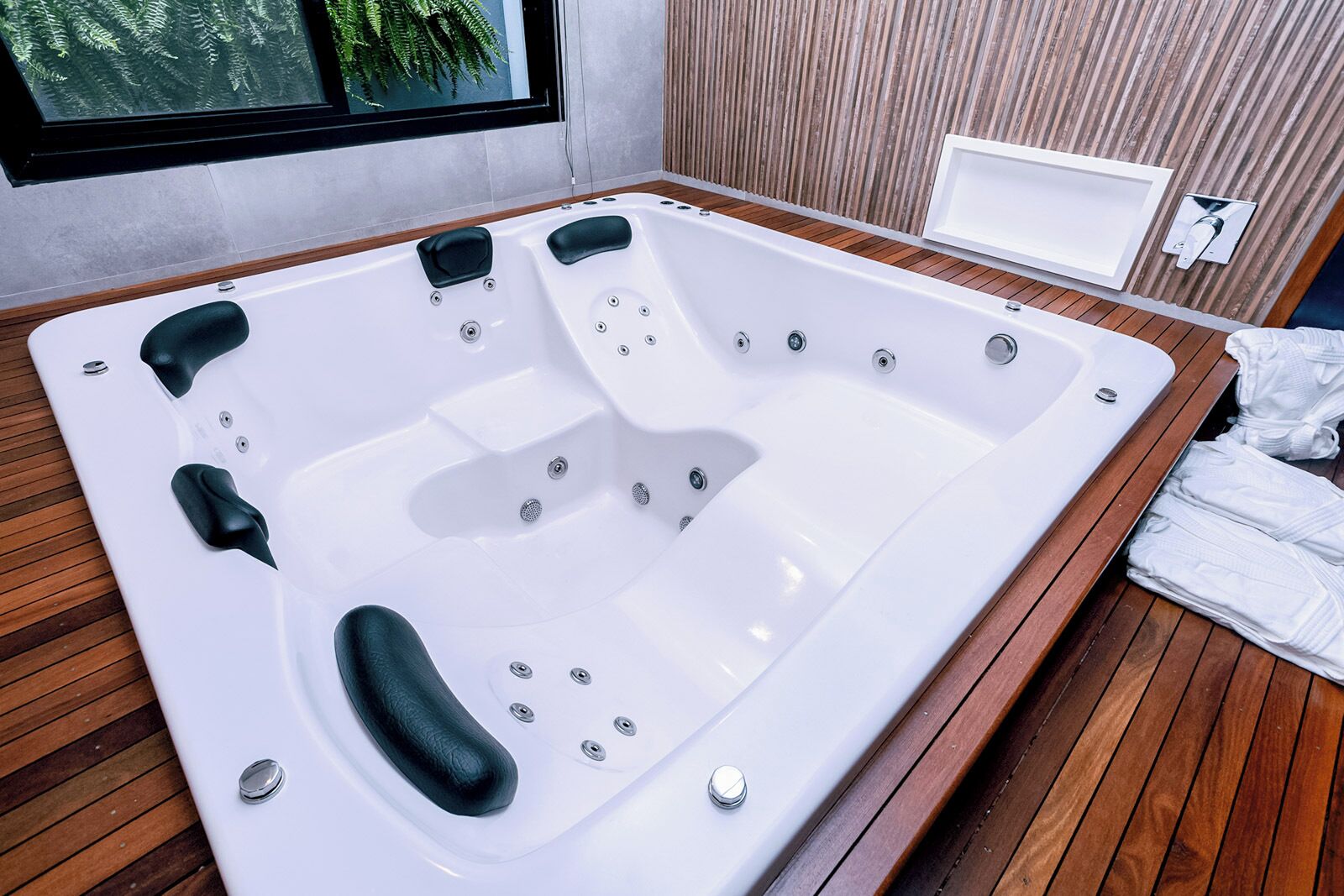 A modern bathroom with a large white bathtub, wooden flooring, and a window with a view of greenery.