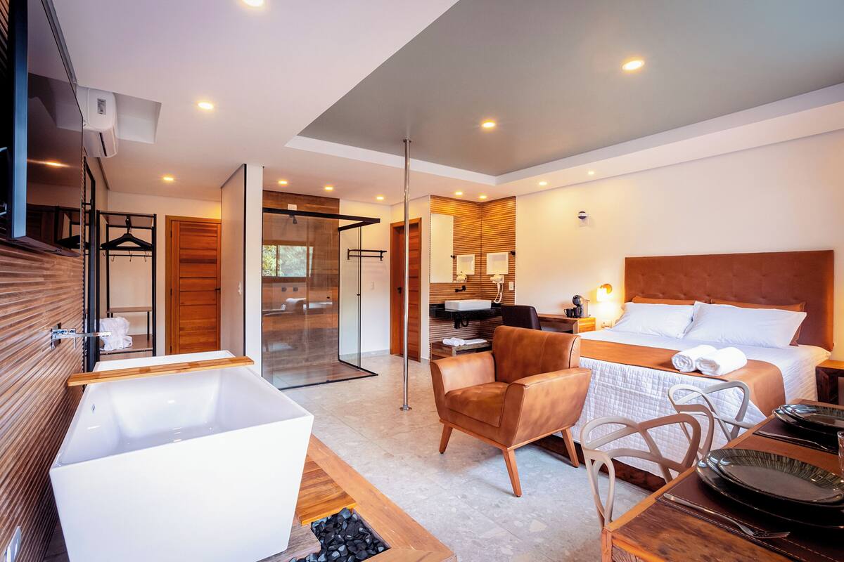 Quarto de hotel moderno com uma banheira grande, uma cadeira de madeira, uma mesa de jantar posta e uma cama grande com cabeceira de madeira.