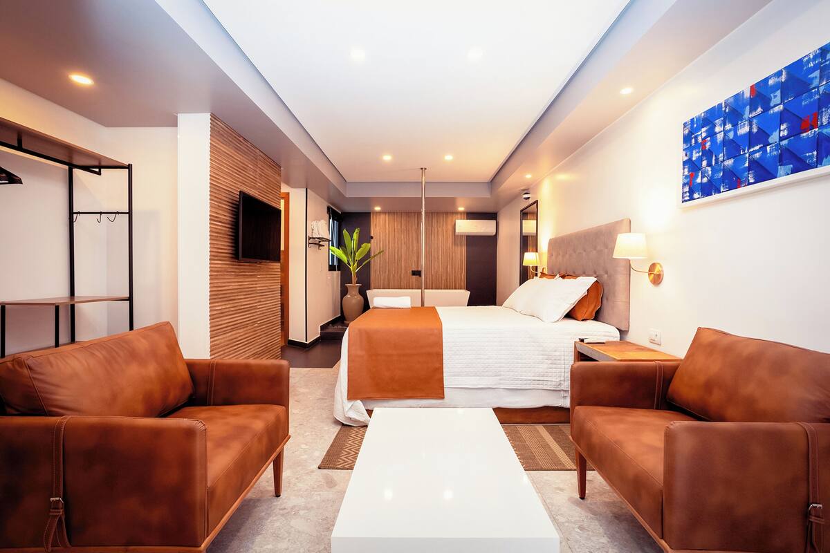 Quarto de hotel moderno com uma cama grande, dois sofás de couro, uma mesa de centro branca e uma janela ampla.