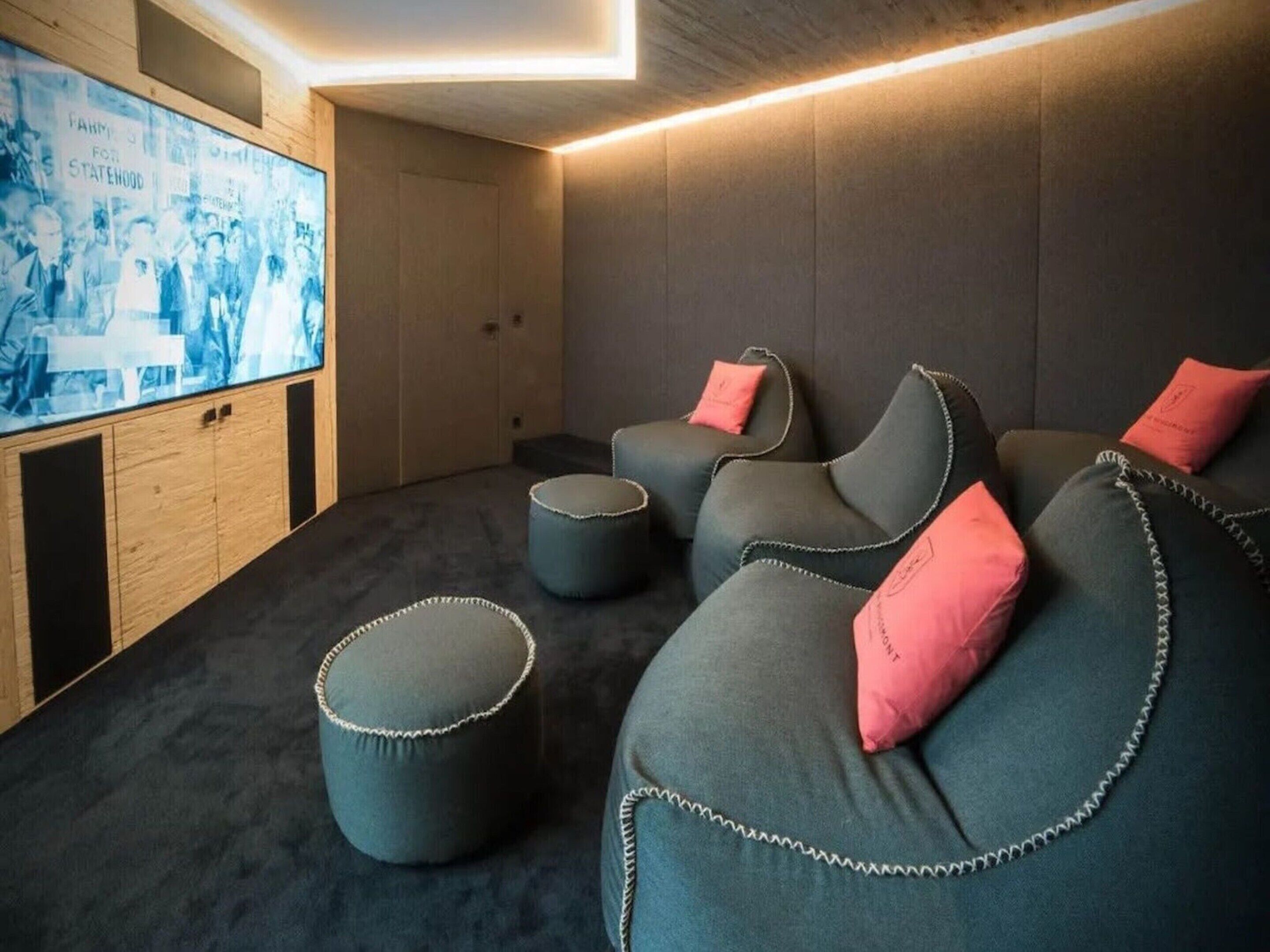 A cozy room with a dark carpet, a large screen, and three grey sofas with pink cushions.