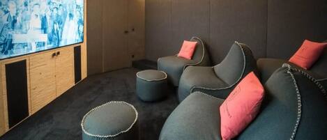 Une pièce chaleureuse avec une moquette sombre, un grand écran et trois canapés gris agrémentés de coussins roses.