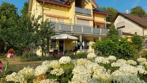 A yellow building with a balcony, surrounded by lush greenery and white flowers.
