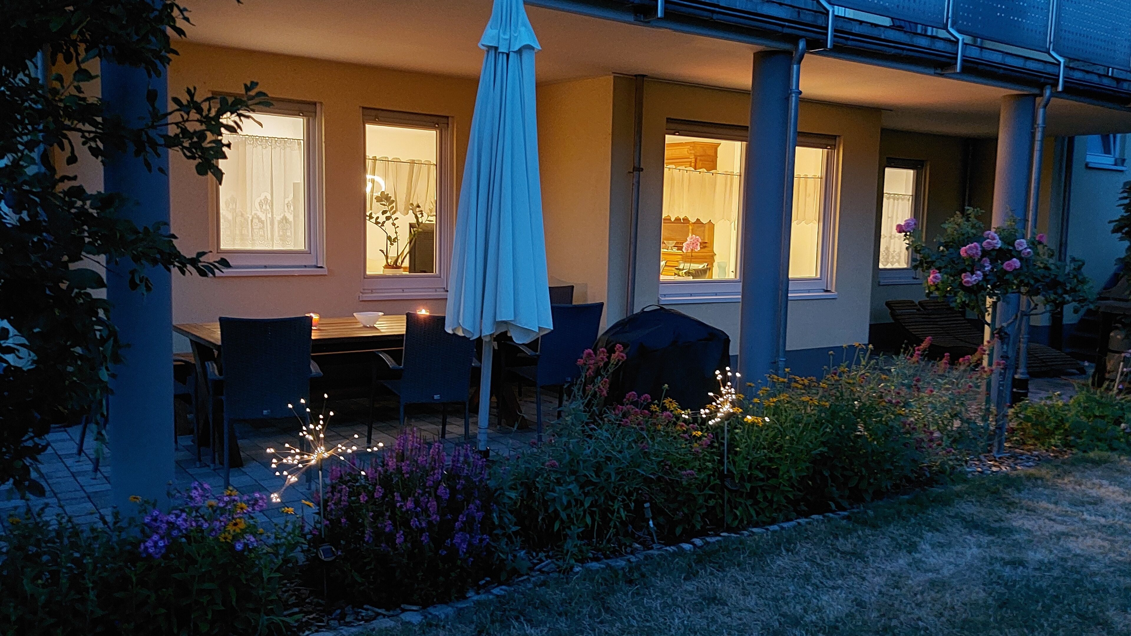 Un espace extérieur aménagé pour les repas, avec des tables et des chaises, un parasol et un jardin fleuri.