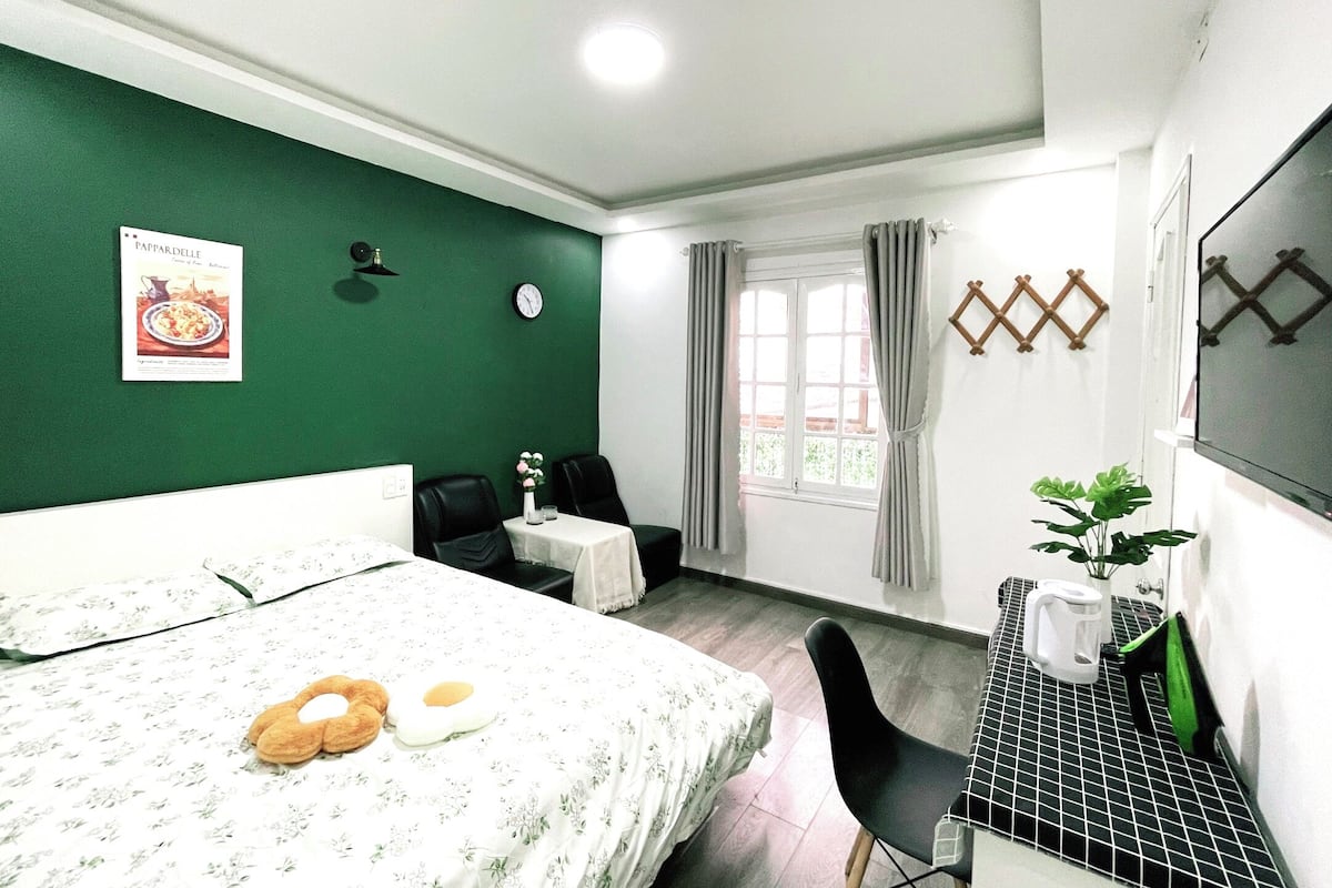 A hotel room with a bed, a small table with a vase, a chair, a TV, and a window with curtains.