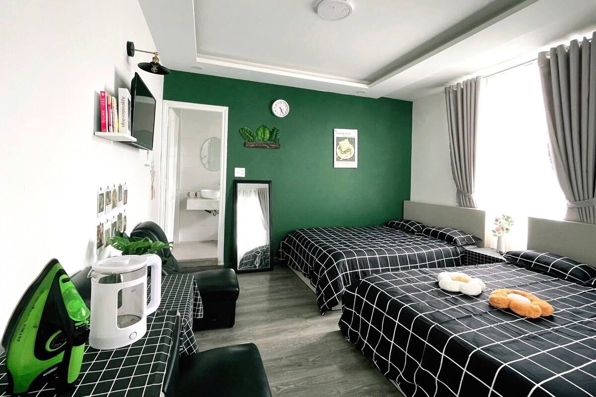 A room with two beds, a desk, a green wall, a window with curtains, and a visible door leading to a bathroom.