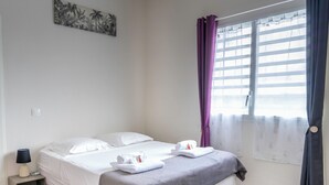 Ein ordentlich bezogenes Bett mit weißen Leinen und einer grauen Decke, ein kleiner Nachttisch mit einer Lampe und ein Fenster mit violetten Vorhängen.