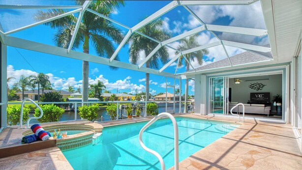 Zona de piscina con agua azul cristalina, una estructura de cristal y vistas a palmeras y casas al fondo.