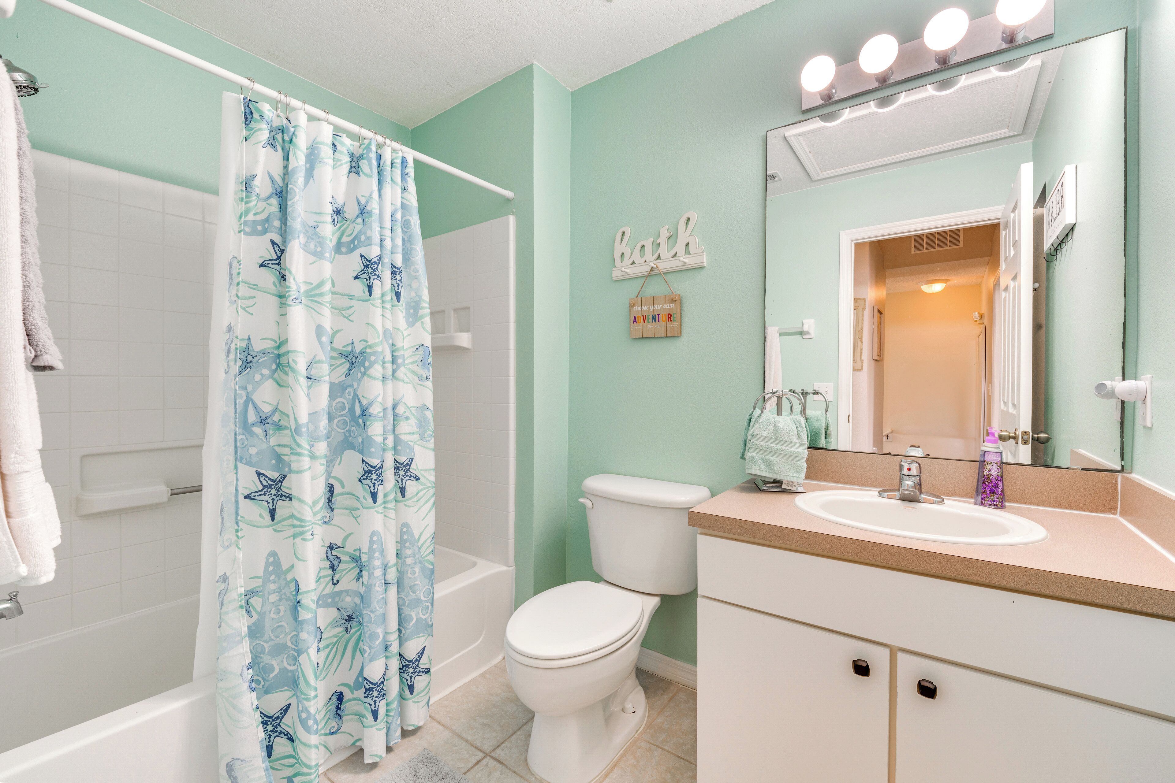A bathroom with a shower curtain, toilet, sink, and a mirror with lights.