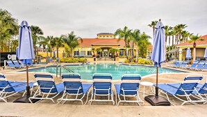 A pool area with blue lounge chairs and umbrellas, surrounded by palm trees and a building with a red roof. - Townhome 7 Mi to Disney Parks: Resort Pool Access! (Kissimmee)