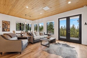 A modern living room with a wooden ceiling, large windows, and a view of trees. - Idyllic 5BD Montana Home w/ private hot tub, fire pit & amazing views! Sleep 8! (Kalispell)
