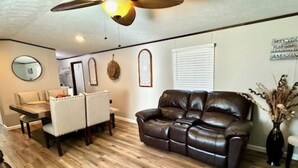 A living room with a brown leather sofa, a dining table with chairs, a ceiling fan, and a wooden floor. - The Rustic Hideaway  A Country Experience Horseback Riding and Fishing  (Cantonment)