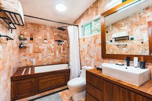 Une salle de bain avec un mur en marbre, un meuble-lavabo en bois, un lavabo blanc, une baignoire et des toilettes.