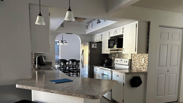 A modern kitchen with a central island, bar stools, and a dining area in the background. - Spacious 3BR home with Heated Pool & WiFi! Great spot near beaches & shopping! (Port Charlotte)