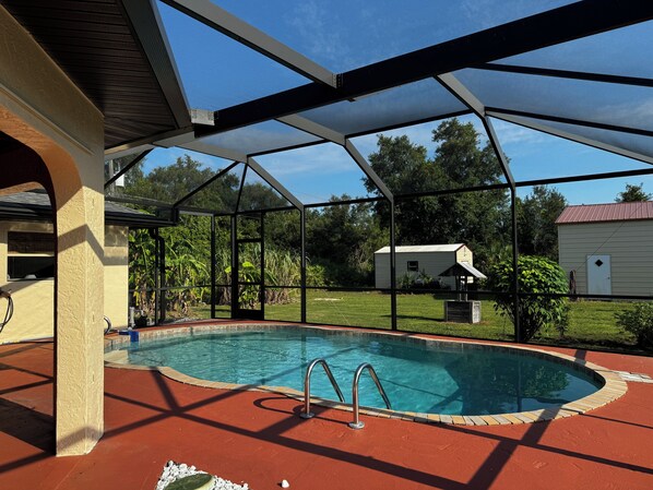 A covered swimming pool with a red deck, surrounded by a metal fence and a view of a house and trees in the background. - Spacious 3BR home with Heated Pool & WiFi! Great spot near beaches & shopping! (Port Charlotte)