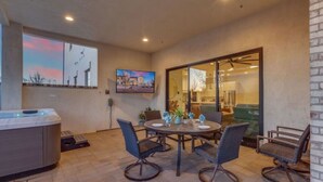 A spacious patio area with a round table and chairs, a hot tub, and a large window with a cityscape view. - Desert Haven Retreat Hot Tub Fire Pit Near Zion (Hurricane)
