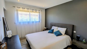 Un dormitorio moderno con una cama grande, una mesita de noche, cortinas translúcidas en la ventana y vistas al verdor exterior.