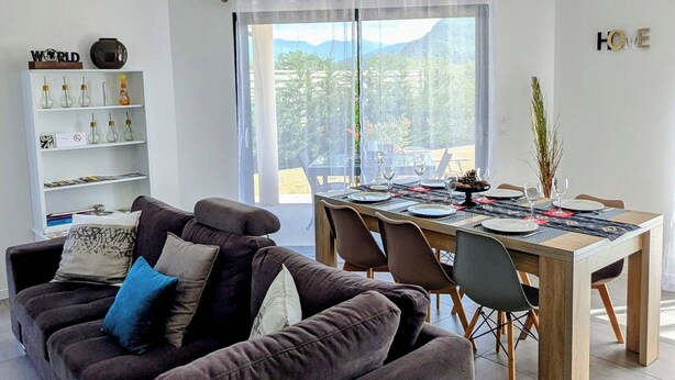 Una sala moderna con un sofá marrón oscuro, una mesa de comedor para cuatro personas y una estantería con adornos.