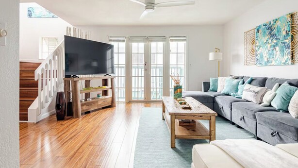 Una sala de estar con un televisor sobre un soporte de madera, un sofá gris con cojines azul turquesa, una mesa de centro de madera y una escalera con barandilla blanca.