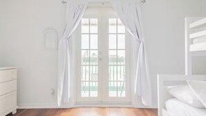 Una habitación luminosa con piso de madera, paredes blancas, un tocador blanco y una ventana con cortinas blancas.