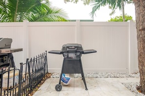 Una parrilla con un saco de carbón en un patio trasero con una valla blanca y un árbol.