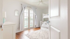 Una habitación luminosa y blanca con piso de madera, ventilador de techo, ventana con cortinas, una cama litera y una pequeña alfombra.