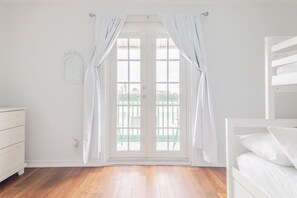 Una habitación con piso de madera, paredes blancas, un aparador blanco y una ventana con cortinas blancas.